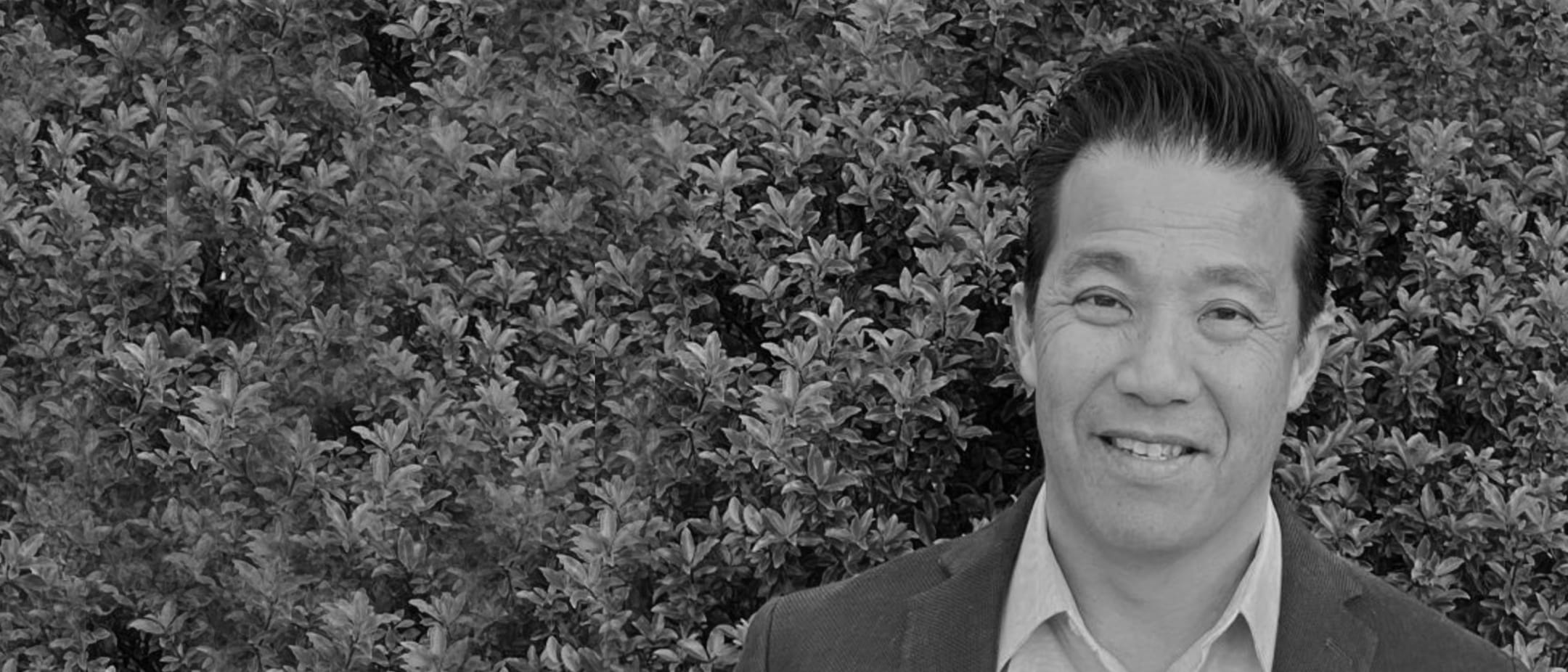 Black-and-white photo of a person wearing a blazer and light shirt, smiling, with a dense leafy hedge in the background.