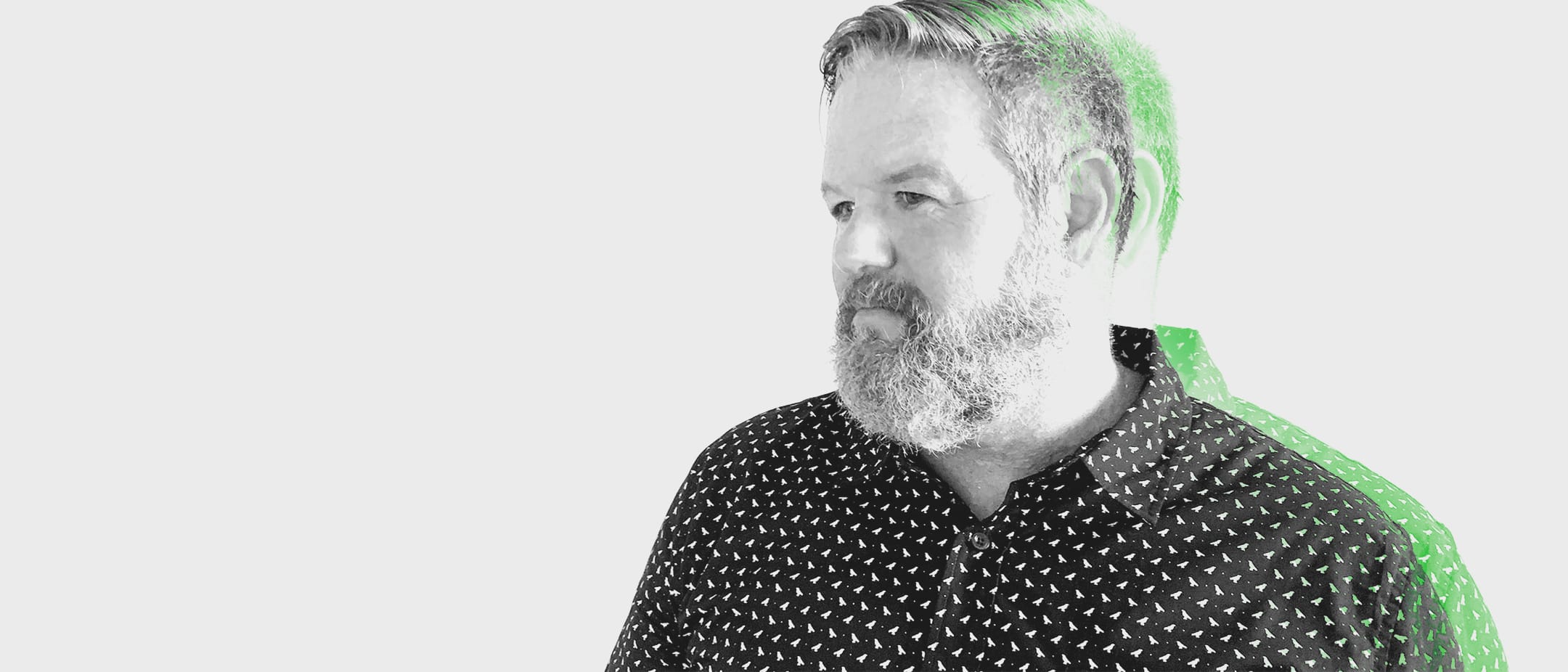 Bearded person with short hair, wearing a dark shirt with small white triangles, looking to the left; green ghosted double exposure behind.