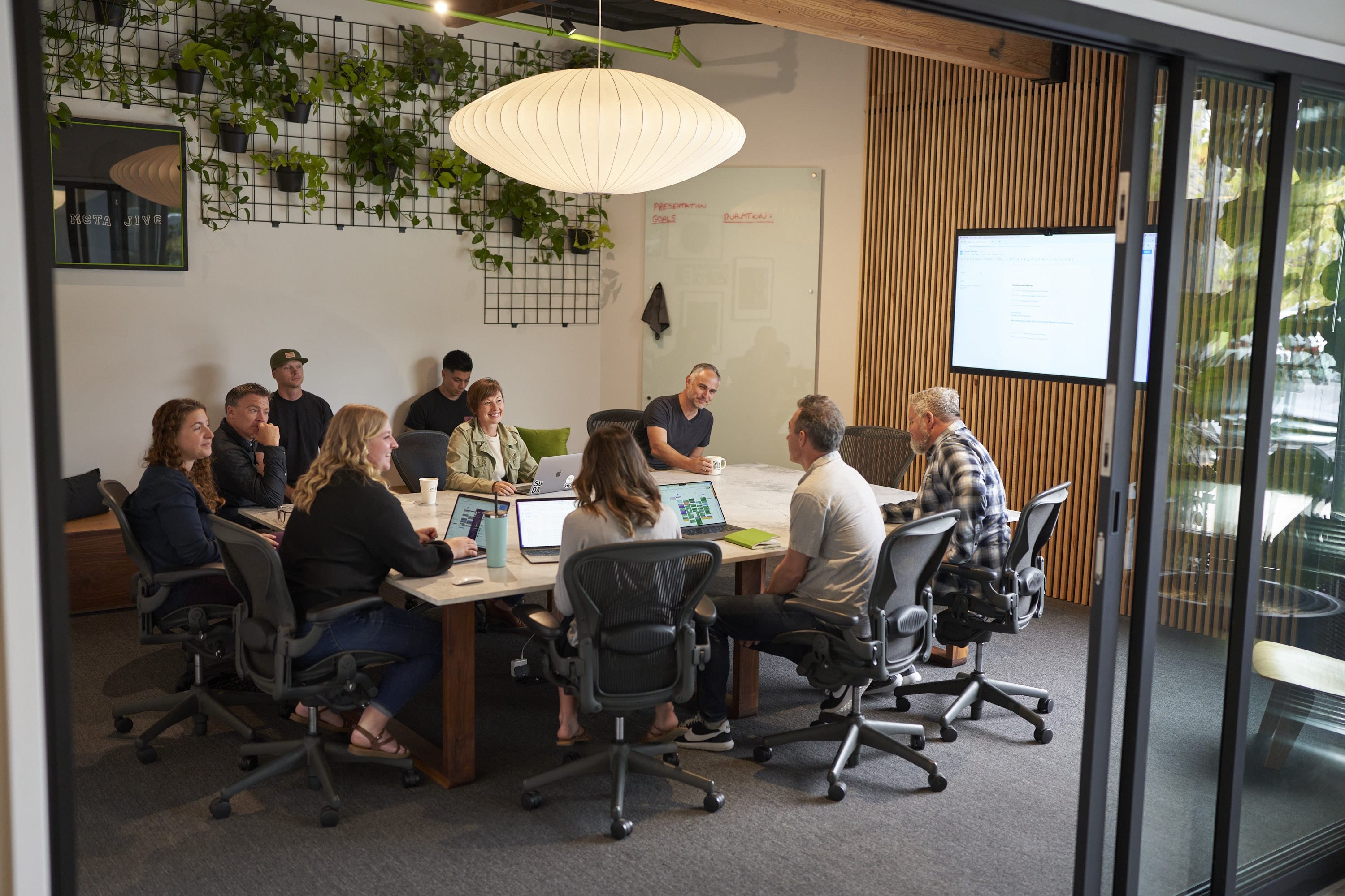 Group of people sit around a large conference table in a modern office; laptops open, whiteboard, screen, hanging lamp, and leafy wall decor.