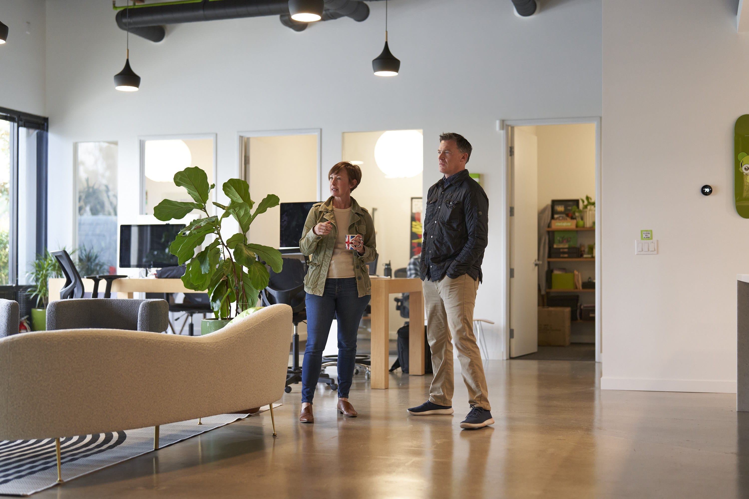 Two people chat in a modern open-plan office lounge with a large potted plant, beige sofa, and desks in the background; one holds a mug.