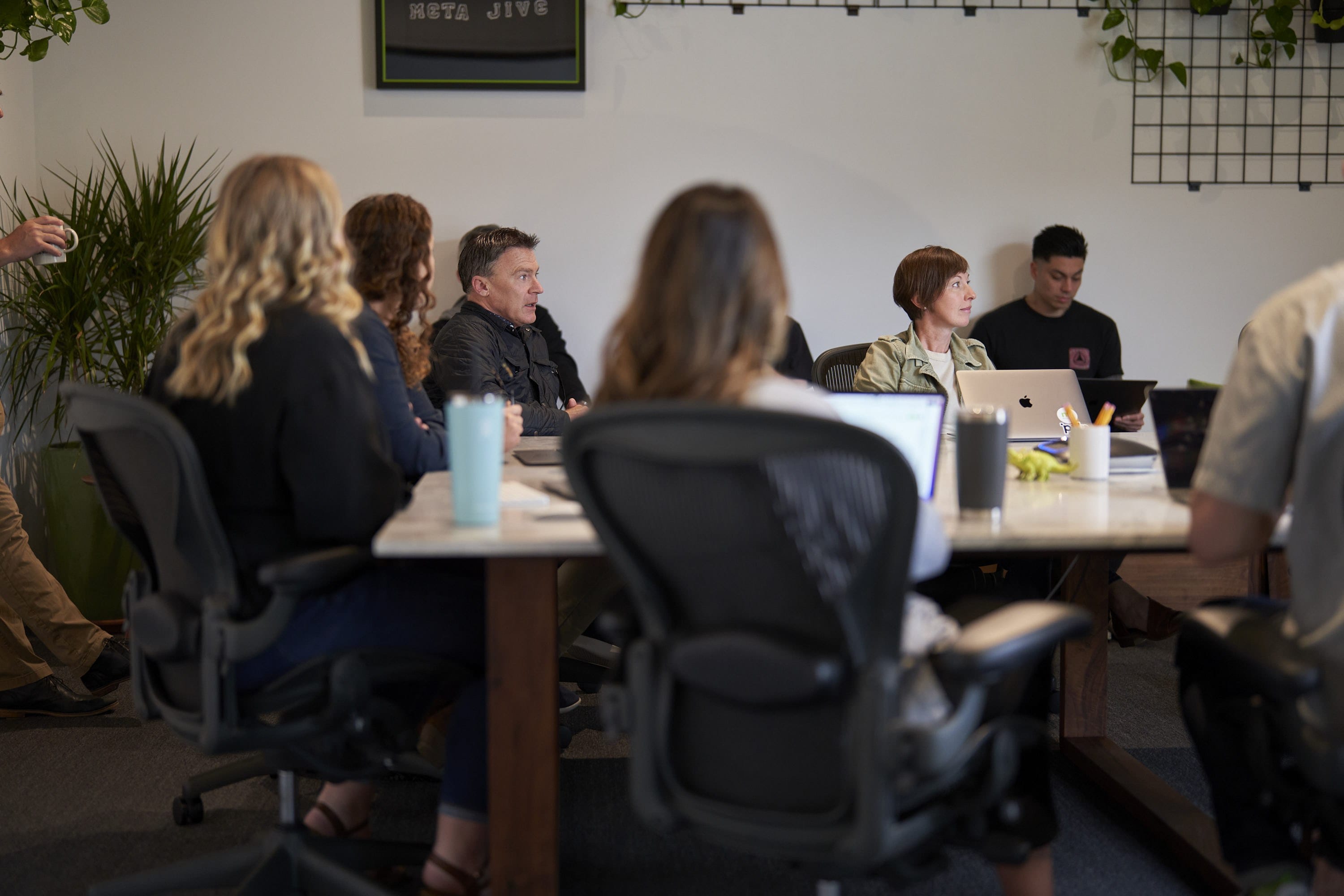A diverse group seated around a rectangular conference table with laptops, cups, and plants in a bright office during a meeting.
