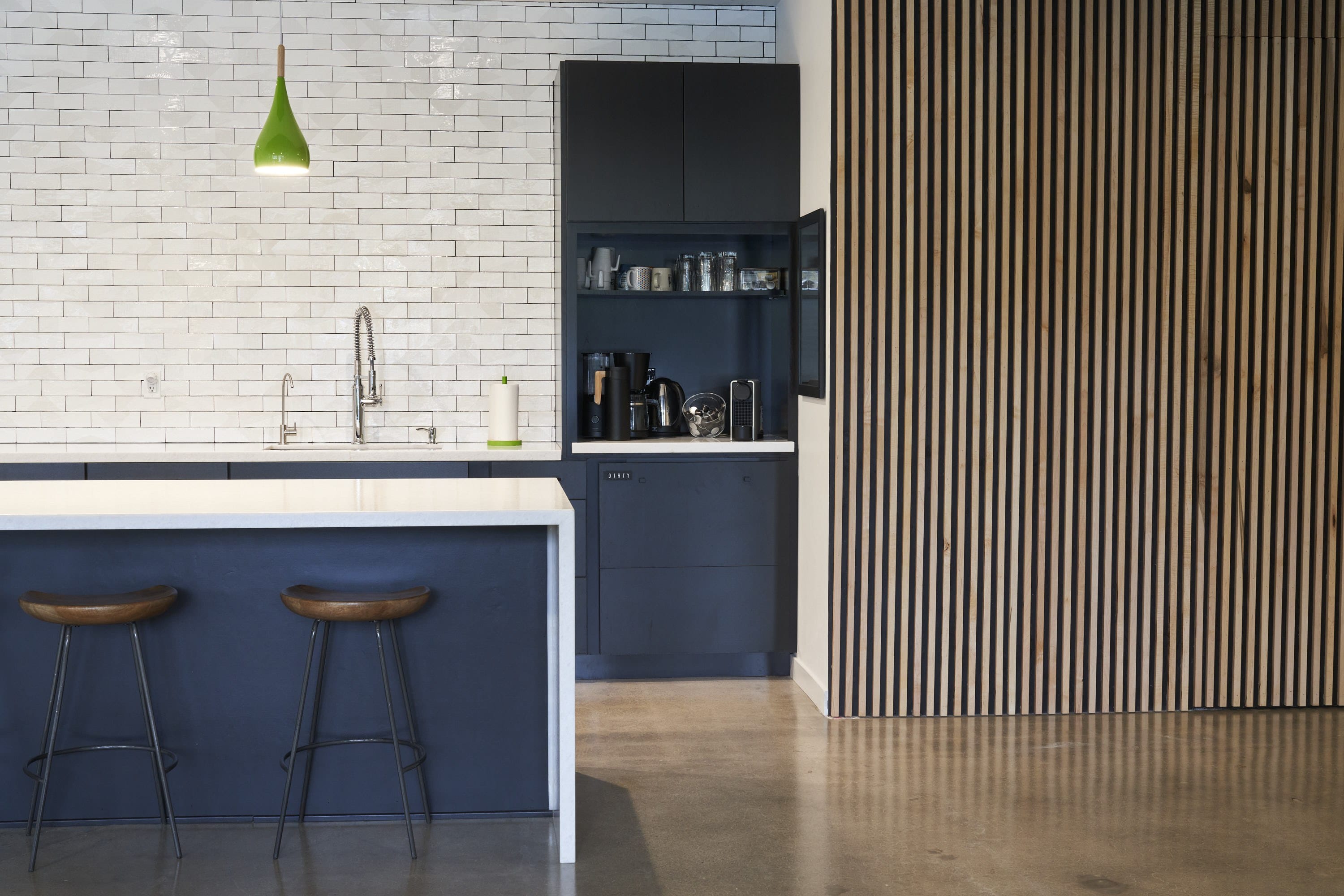 Modern kitchen with white subway tile, dark blue cabinets, a white island with two wooden stools, a green pendant light, and a vertical slat wood wall.
