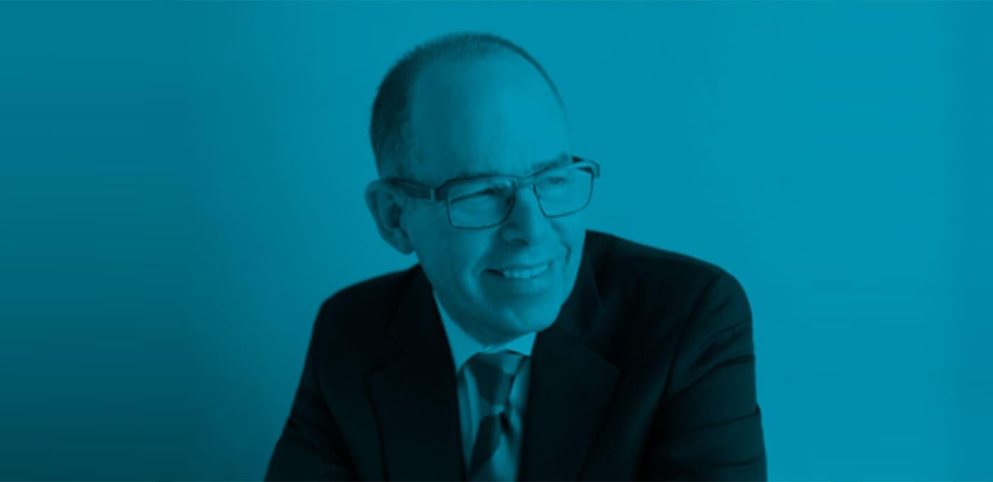 Person wearing glasses and a suit with tie, smiling slightly and looking to the right against a teal-blue background.