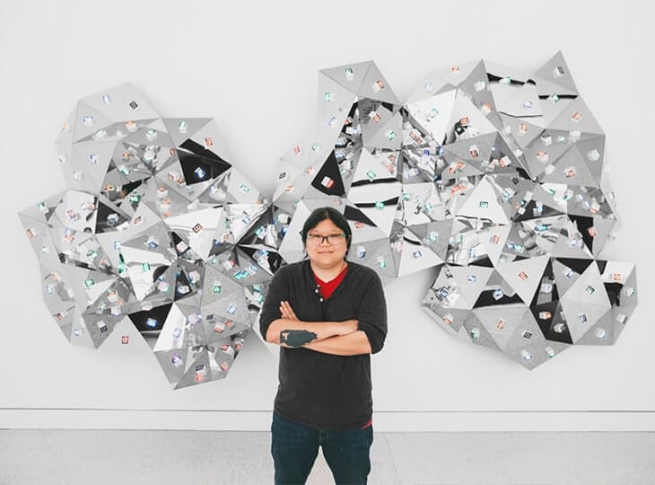 A person with glasses and dark clothing stands with arms crossed in front of a wall sculpture made of silver triangular panels covered with small stickers.