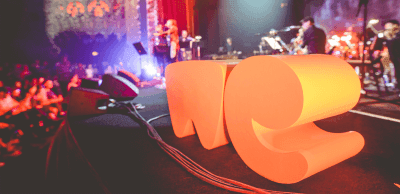 Stage with a live band performing; glowing orange letters spelling LOVE in the foreground, colorful lights and cheering crowd in the background.