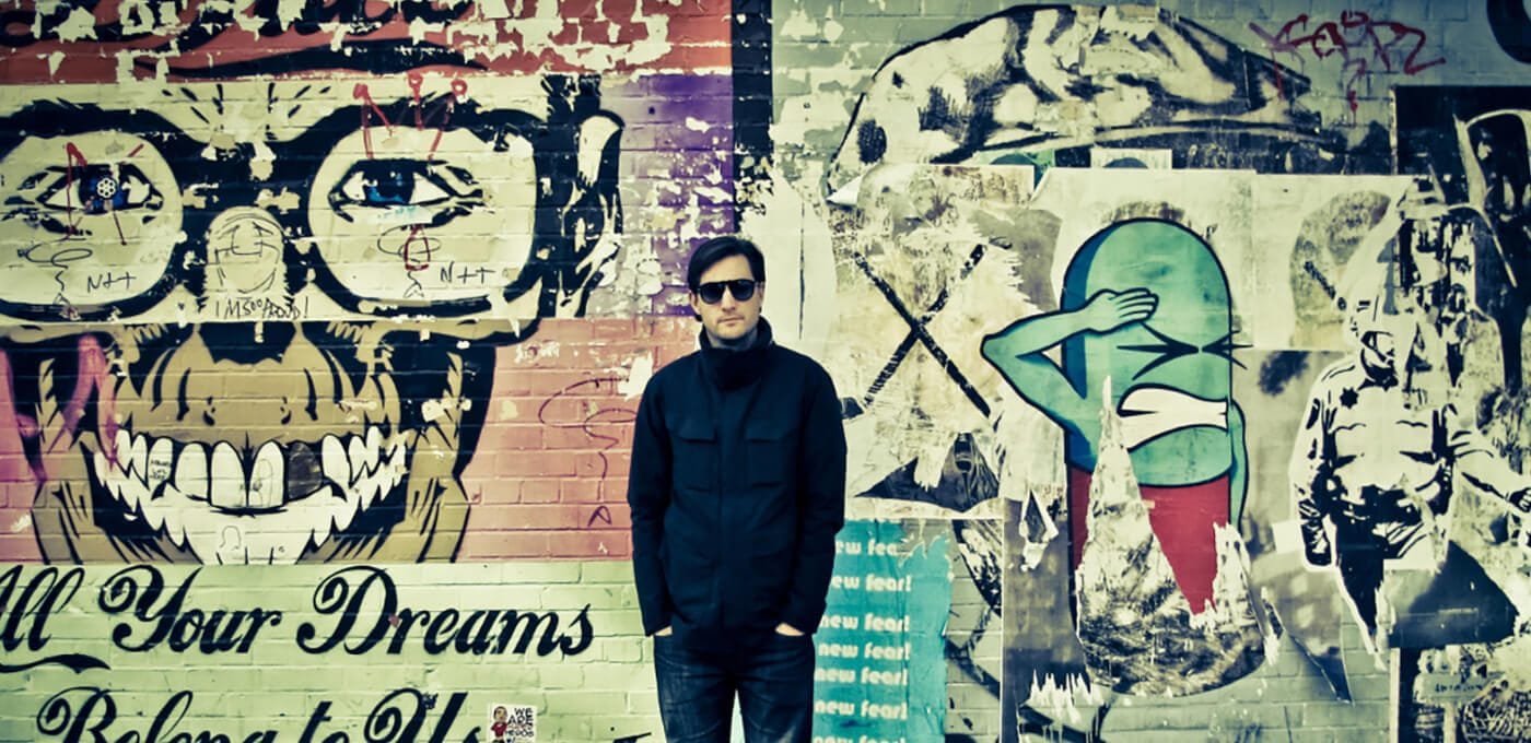 Person in a dark jacket and sunglasses stands before a colorful graffiti mural with a large skull wearing glasses and a blue-green cartoon figure.