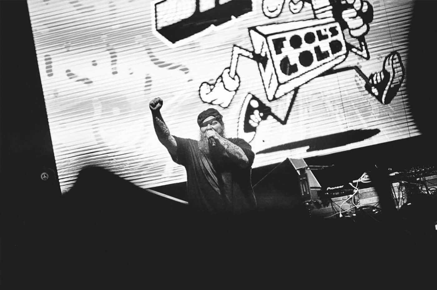 A performer on stage with a microphone, fist raised, tattooed arm visible, in front of a large black-and-white cartoon backdrop.