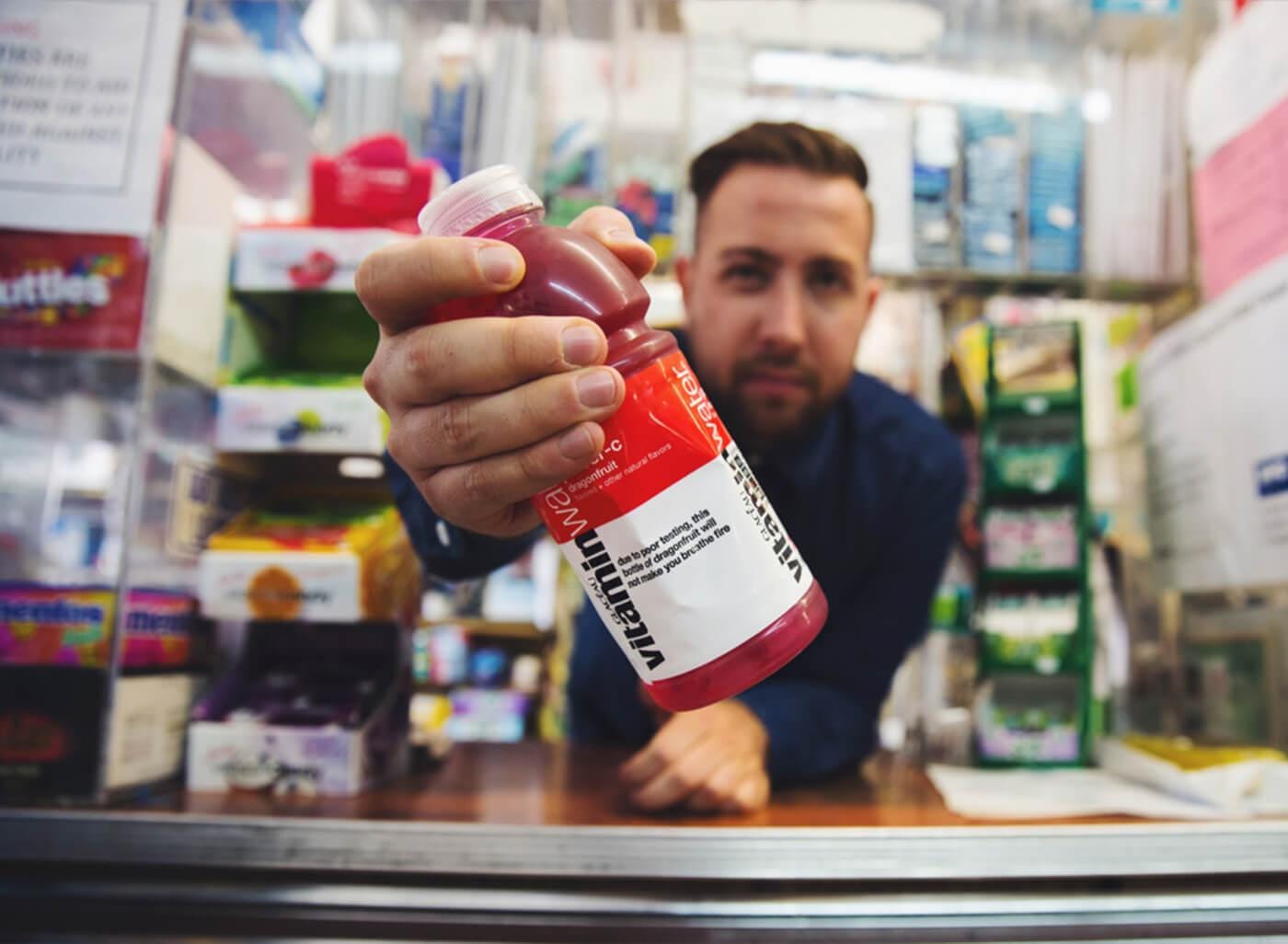 A person at a shop counter extends a pink-red bottle with a white cap toward the camera, with blurred shelves and products in the background.