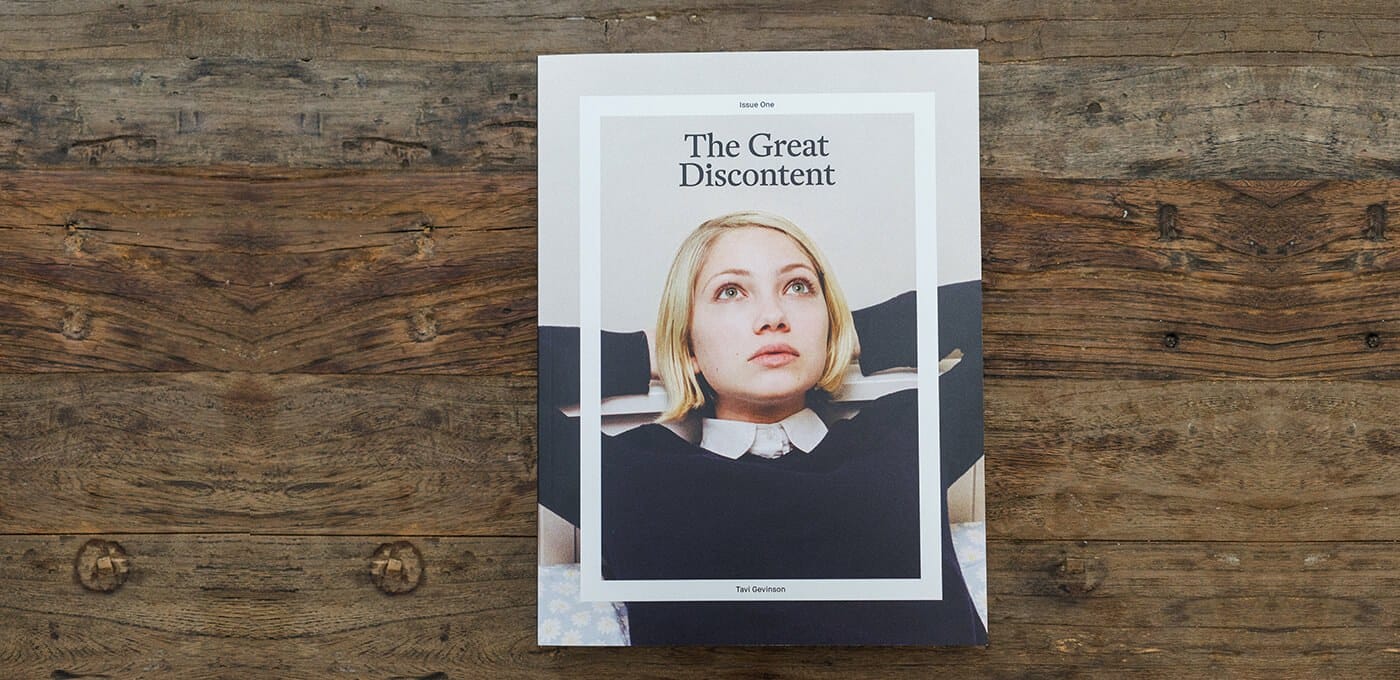 Cover of “The Great Discontent” featuring a person with short blond hair looking upward, framed by a white border, on a wooden table.