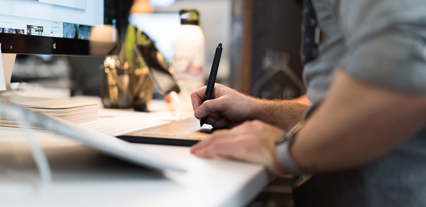 Person drawing on a graphics tablet with a stylus at a desk, in front of a computer monitor, with a keyboard and mug on a bright workspace.