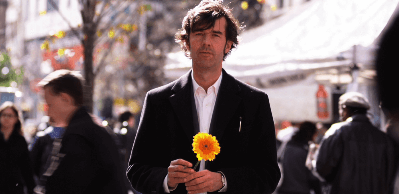A person in a black blazer and white shirt holds a bright yellow flower on a busy outdoor city street, with blurred pedestrians in the background.