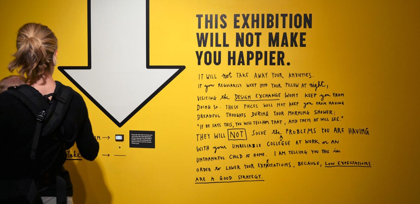Person with a backpack stands before a yellow wall with a large white downward arrow and bold text reading "This exhibition will not make you happier" plus handwritten notes.
