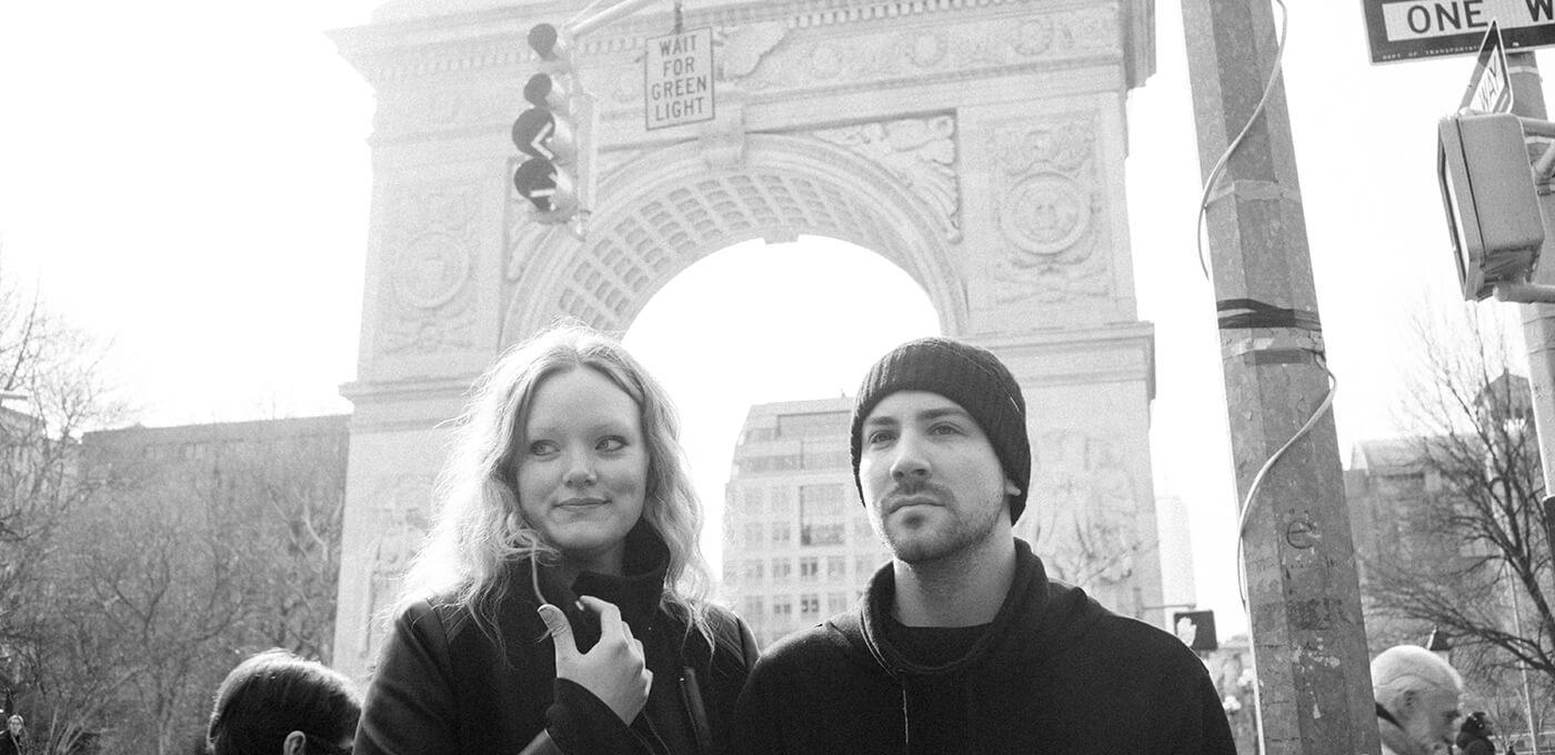 Two people stand in front of a large ornate city arch in a grayscale image; one has light wavy hair, the other wears a knit beanie and dark hoodie, looking ahead.