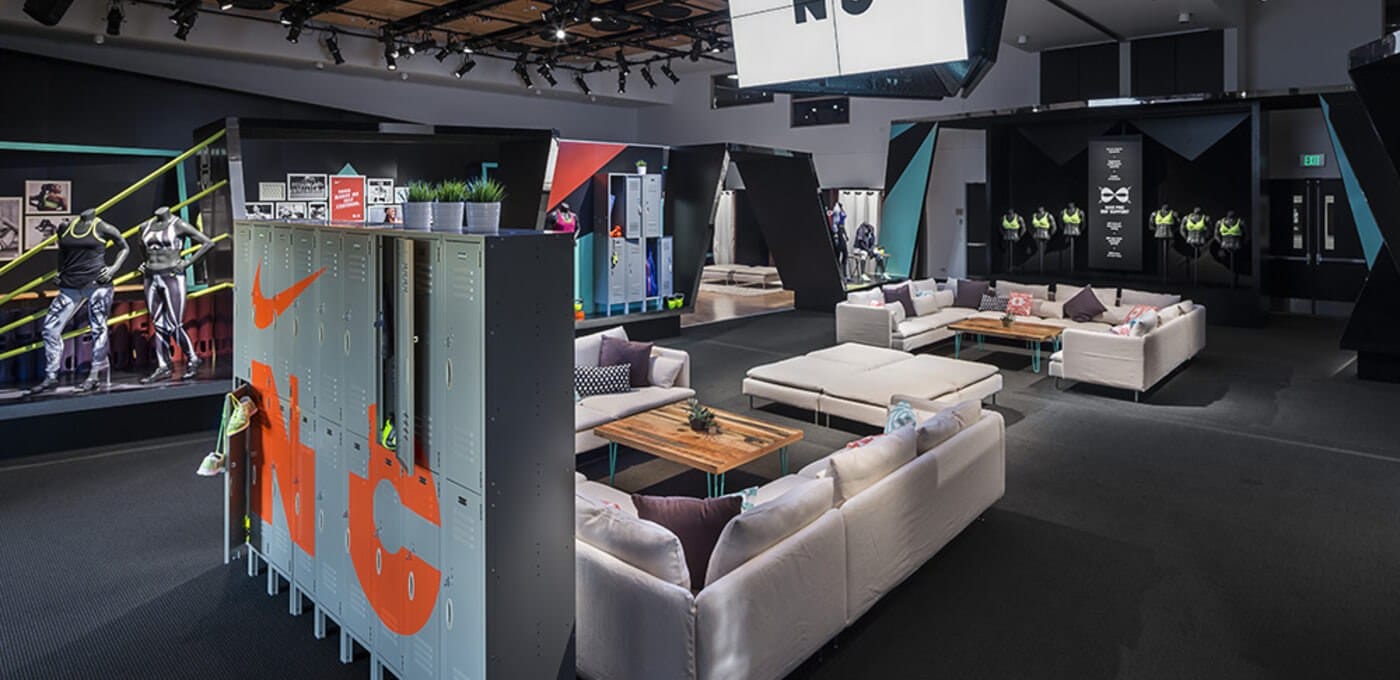 Modern sports showroom with grey lockers featuring an orange Nike swoosh, white sectional sofas, wooden coffee tables, and mannequins near a yellow railing.