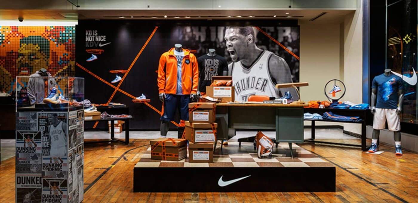 Nike store display featuring a large basketball poster, an orange jacket on a mannequin, stacked boxes, sneakers, jerseys, and a white swoosh on a black platform.