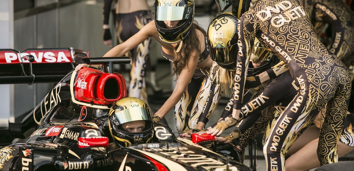 Black-and-gold race car at a pit stop, driver in a gold-visor helmet seated in the cockpit while crew in matching patterned suits push and assist.
