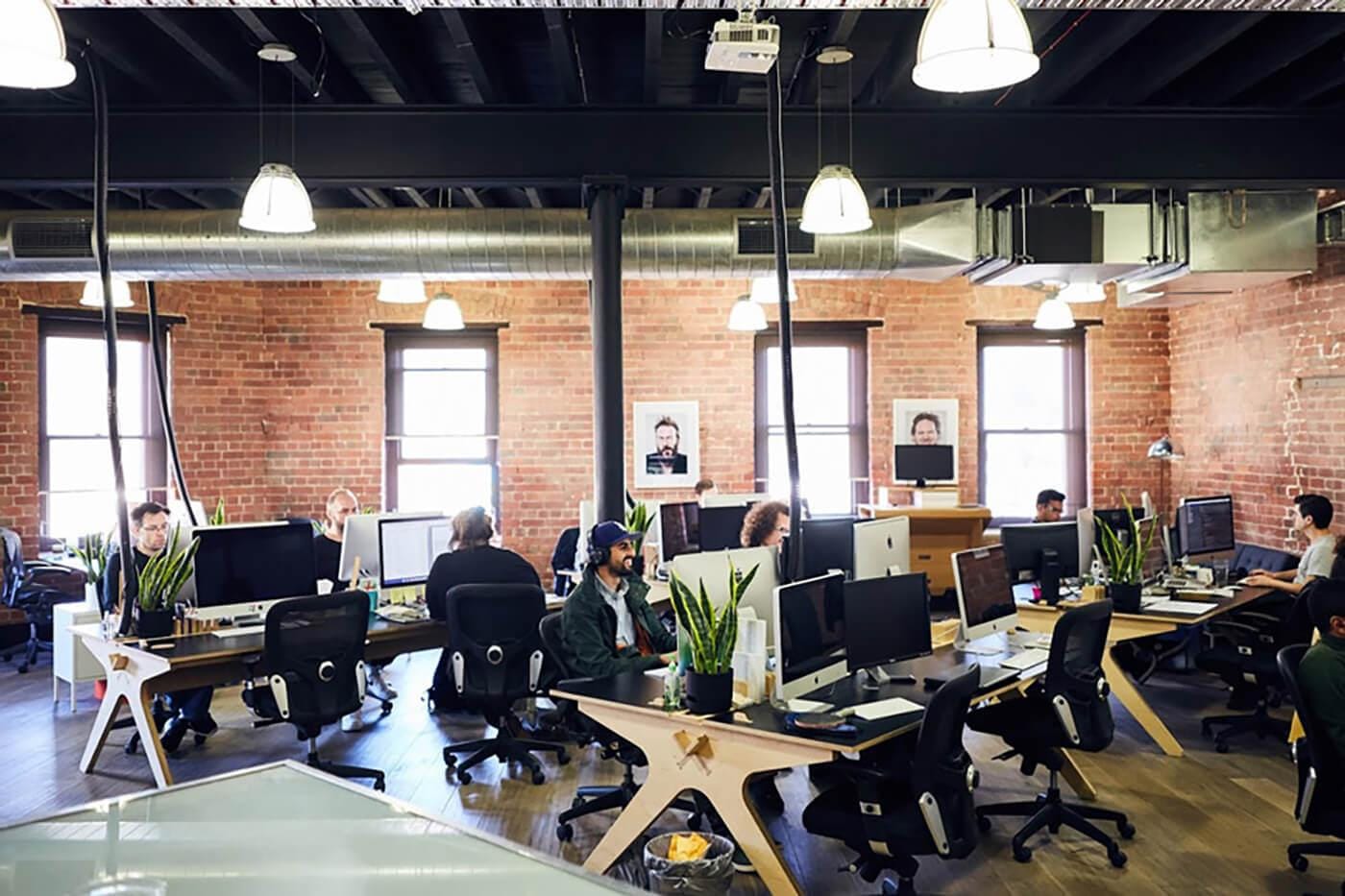 Open-plan office with brick walls, exposed ducts, and hanging lights; rows of desks with large monitors, plants, and workers at computers.