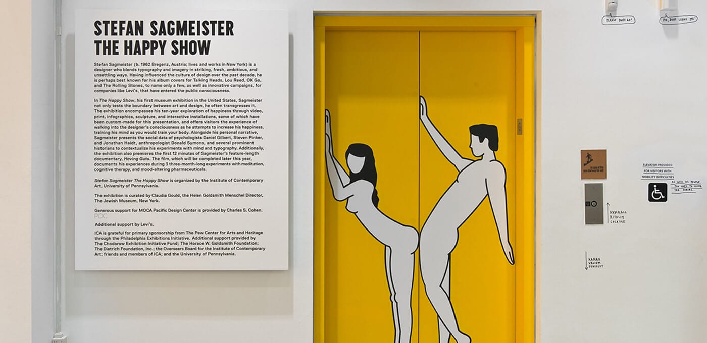 Exhibition wall with a large text panel reading 'STEFAN SAGMEISTER THE HAPPY SHOW' on the left, and a bright yellow door on the right with two white, outline-drawn figures.