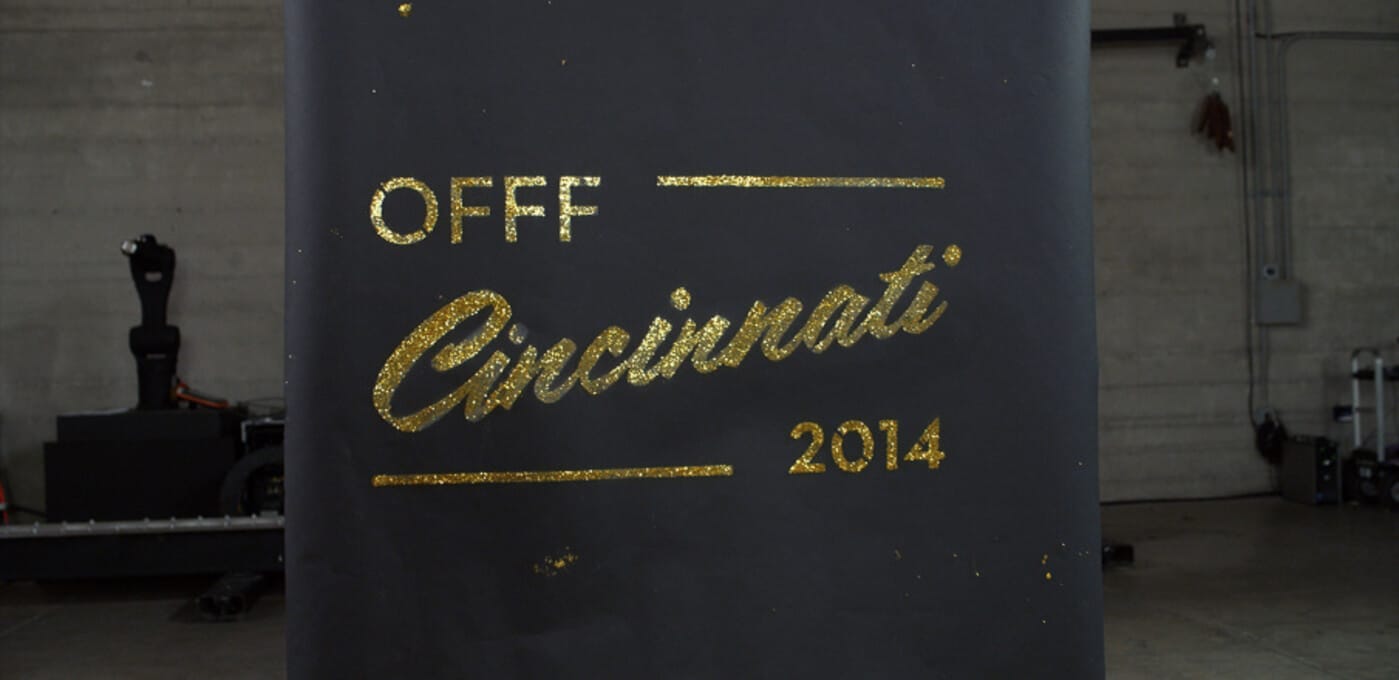 Large dark banner with glittery gold text reading OFF Cincinnati 2014 and decorative lines, set in an industrial warehouse.