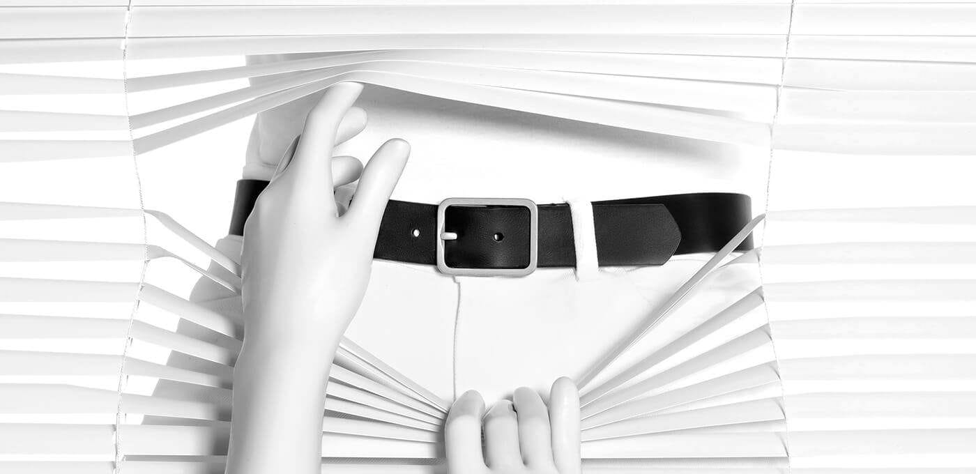 Black-and-white photo of a person behind white blinds, one hand adjusting a black belt around the waist as the blinds open around the torso.