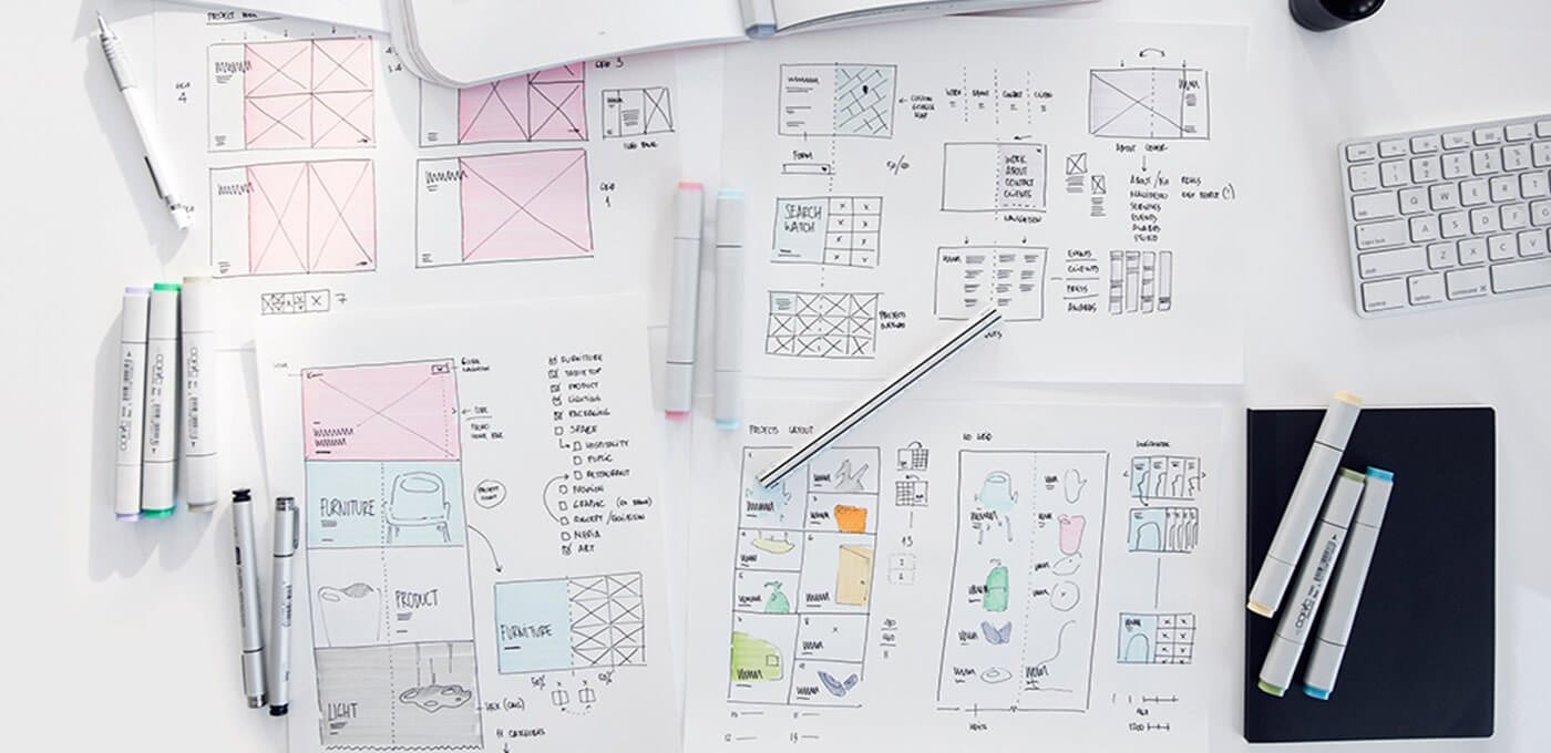 Top-down view of a white desk covered in UI wireframe sketches, colored markers, pens, and a keyboard beside a black notebook.