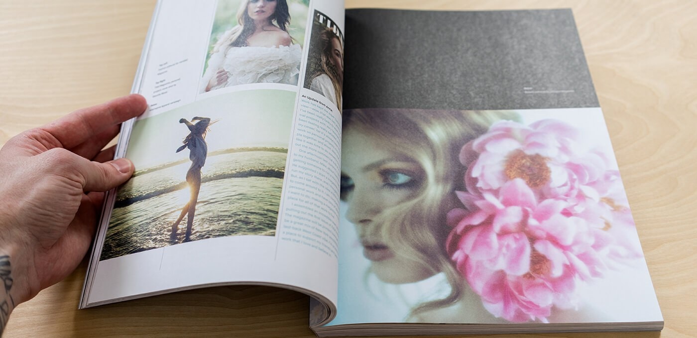 A hand flips through a magazine on a wooden table, showing a beach silhouette on one page and a close-up portrait with pink flowers on the other.