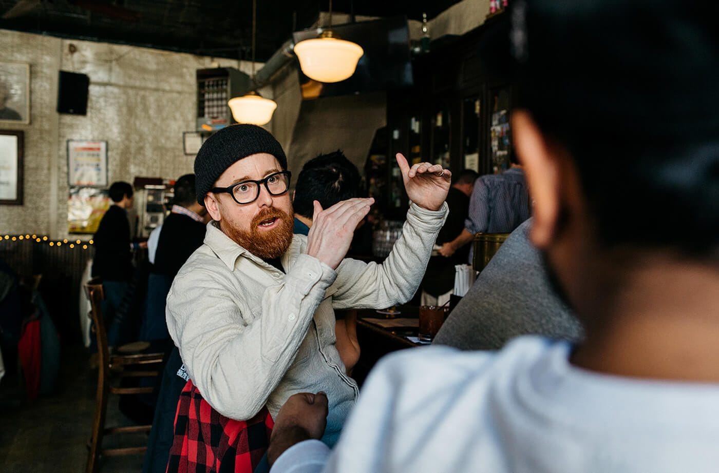 A bearded person with glasses and a black beanie gestures with their hands while seated at a busy cafe or bar, with other patrons in the background.